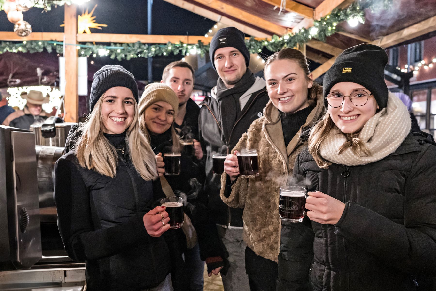 Eine Gruppe von sechs jungen Erwachsenen in Winterkleidung lächelt und hält Tassen mit heißen Getränken auf einem mit Girlanden und Lichterketten geschmückten Weihnachtsmarkt.
